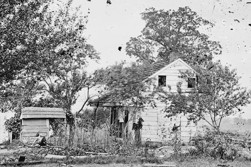 A person walks beside a small house that has a small shed behind it.