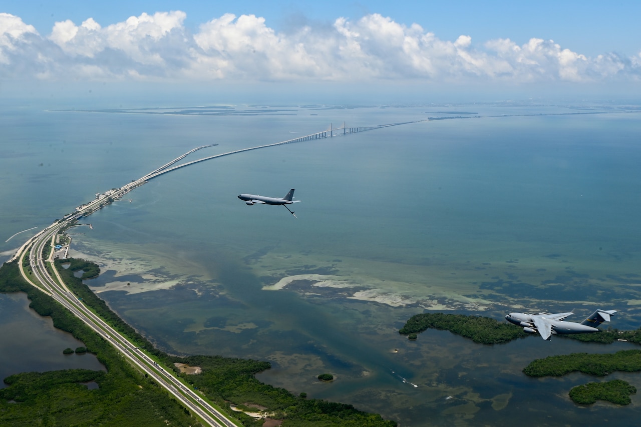 Two planes fly over a long bridge.