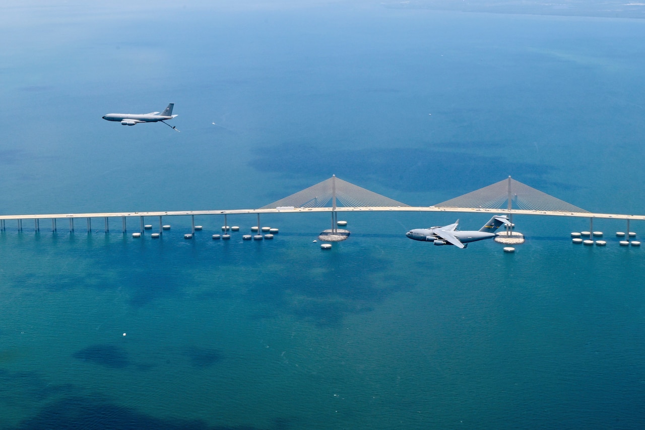 An aircraft releasing its boom travels in front of another aircraft as they fly over a bridge in body of water.
