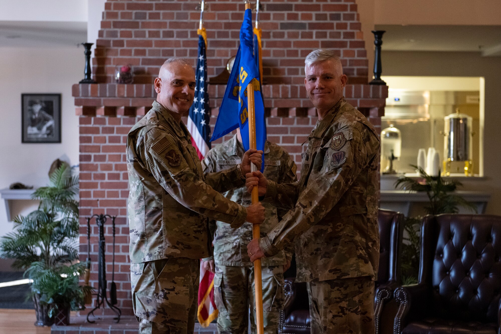 A photo of two military-uniformed men holding a guide on - a wooden post with a flag on the top, in the middle of the two of them, both grasping the pole.