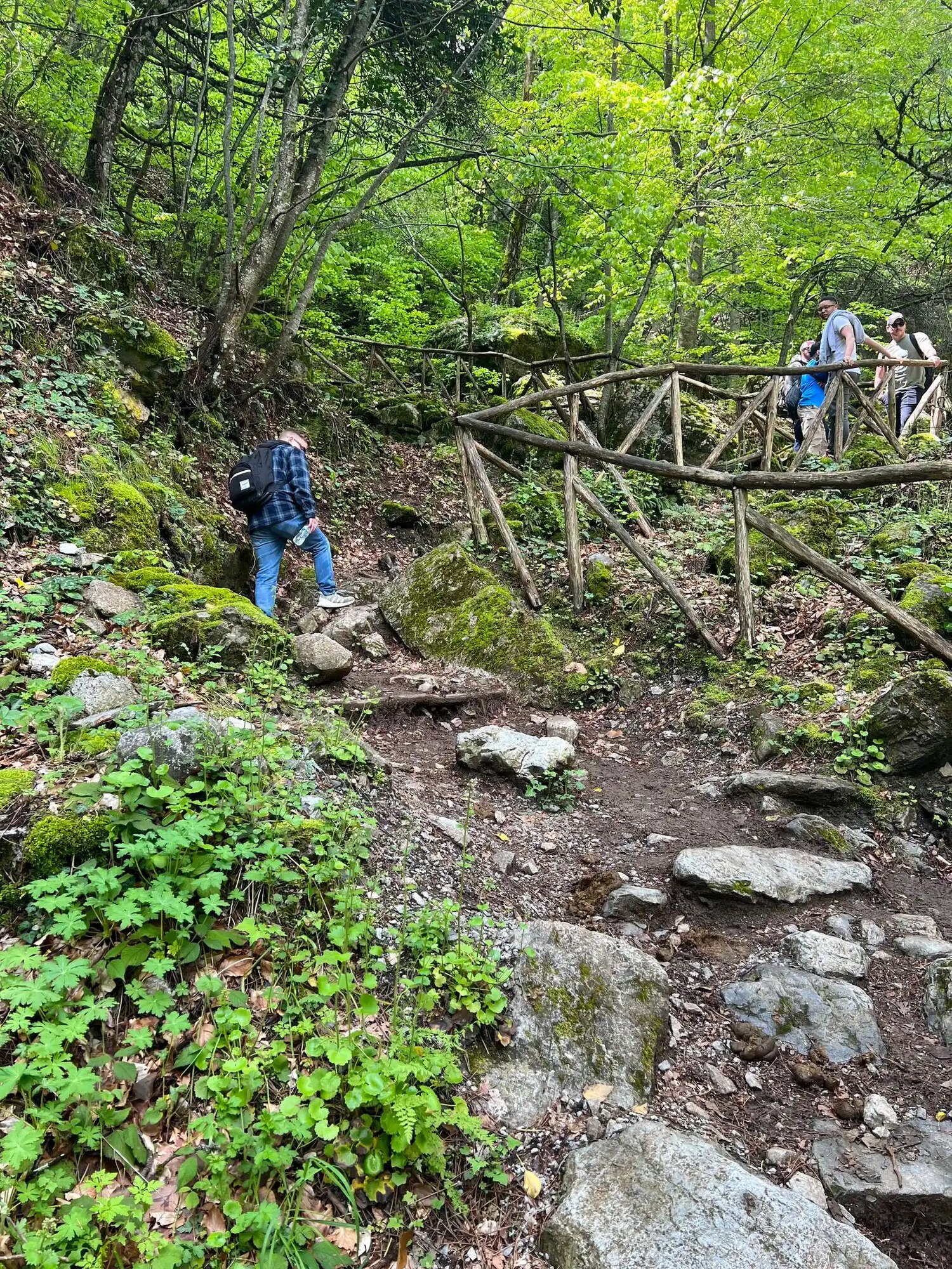 A man climbs up a rocky trail next to a wooden rail while a group of three people wait for him farther along.