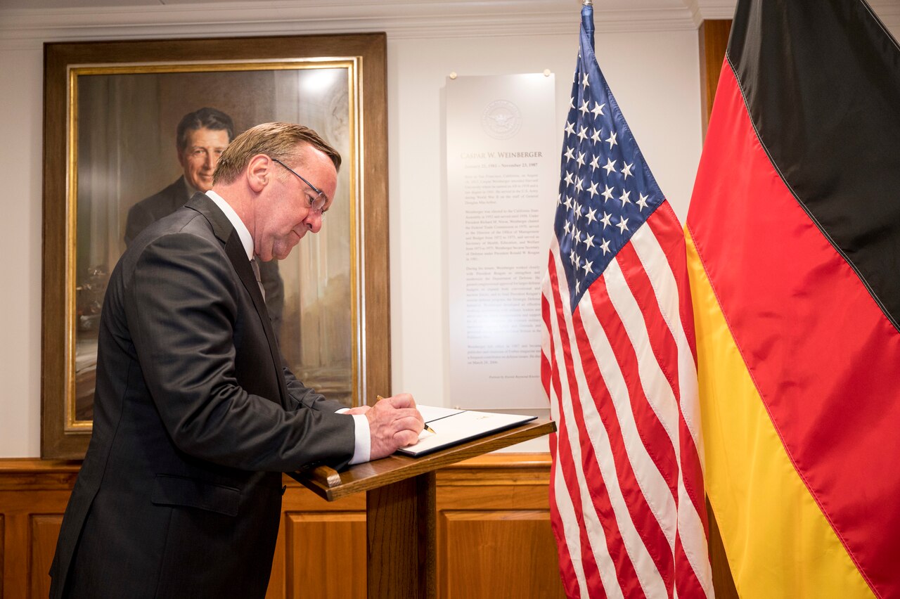A man in a suit signs a book while standing next to U.S. and German flags.