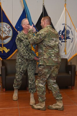 Photo is of a two men in military uniform with one man passing a rolled up flag to another