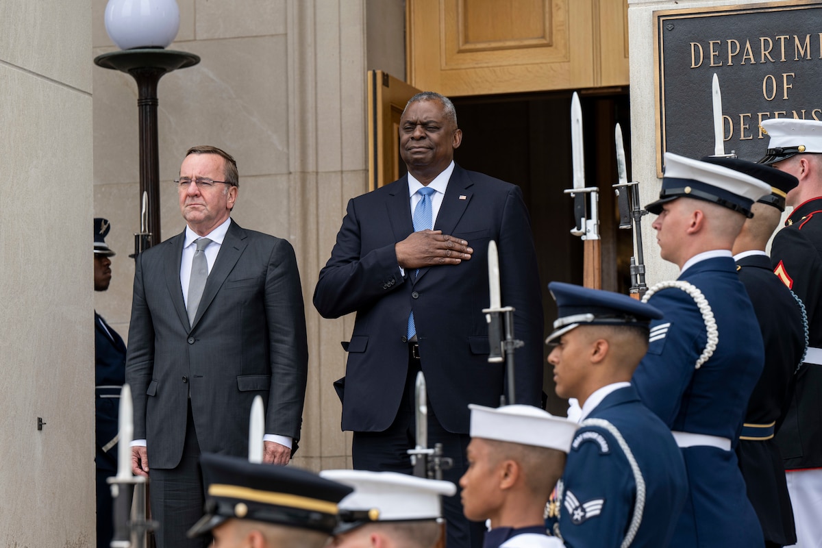 Two men in suits stand next to one another; they’re flanked by a military ceremonial guard.