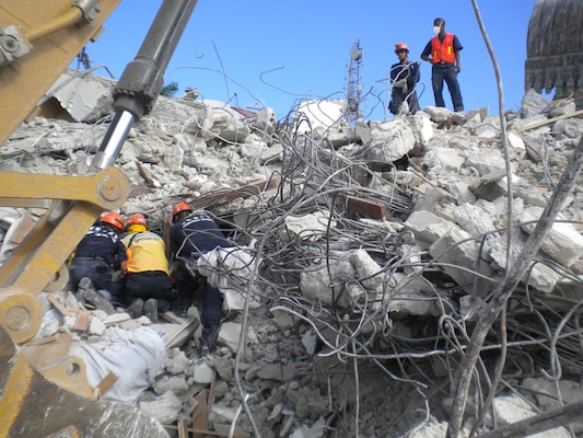 Rescuers search through the rubble of the Hotel Montana. Two men stand atop of the debris while three others sift through the rubble below them.