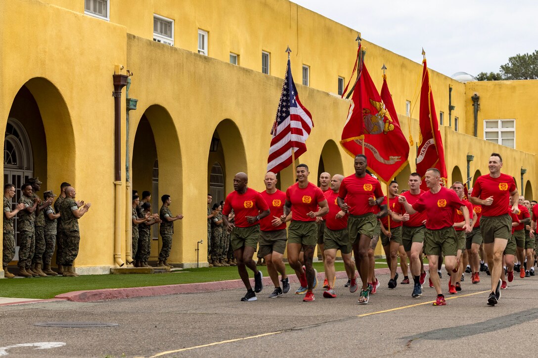 Marines run in formation in the street as fellow service members watch and applaud from the sidewalk.