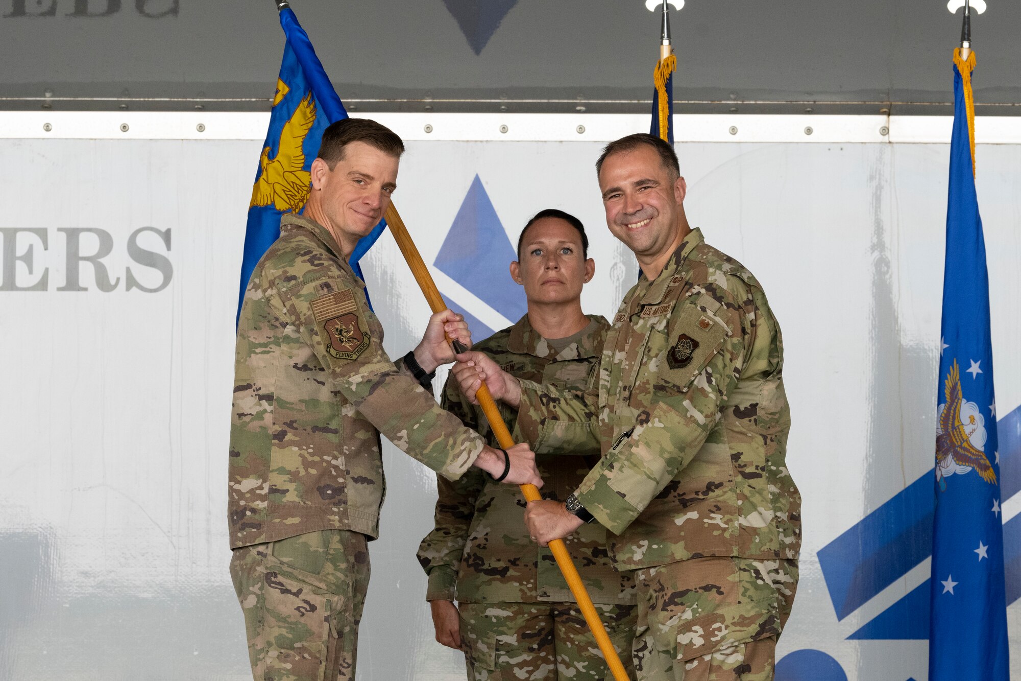 A photo of two men in U.S. Military uniform grasping a guidon between each other, smiling at the camera.