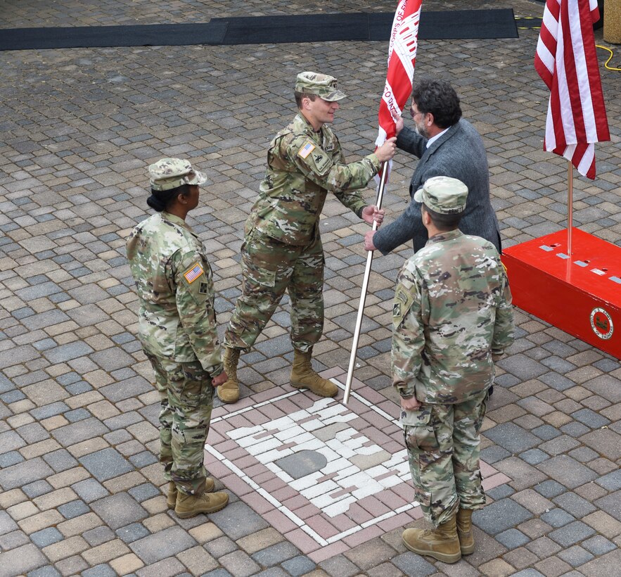 Three soldiers and a civilian stand in a square formation, as a soldier passes a red and white flag to the civilian.