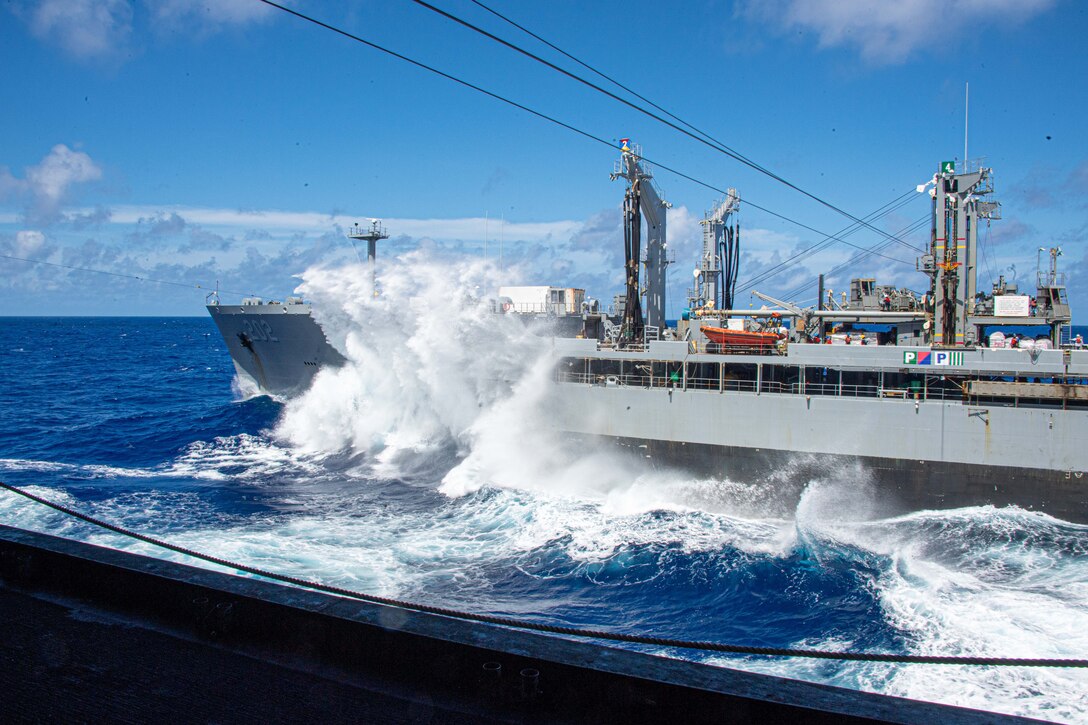 A large wave strikes the side of a ship during a replenishment.