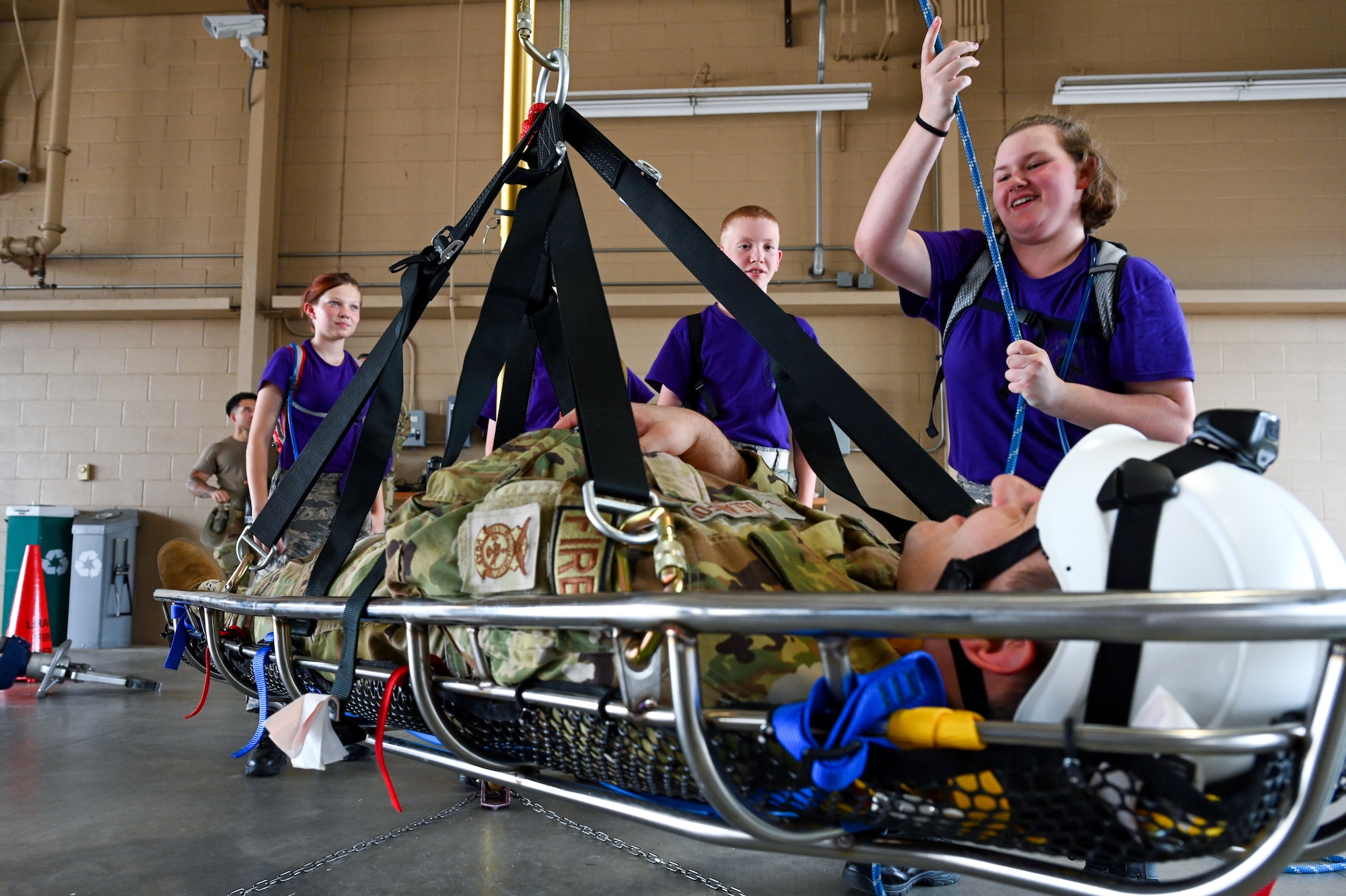 A Civil Air Patrol Cadet wearing a purple shirt lifts up an Airman in a rescue tripod. The Airman is wearing OCPs.