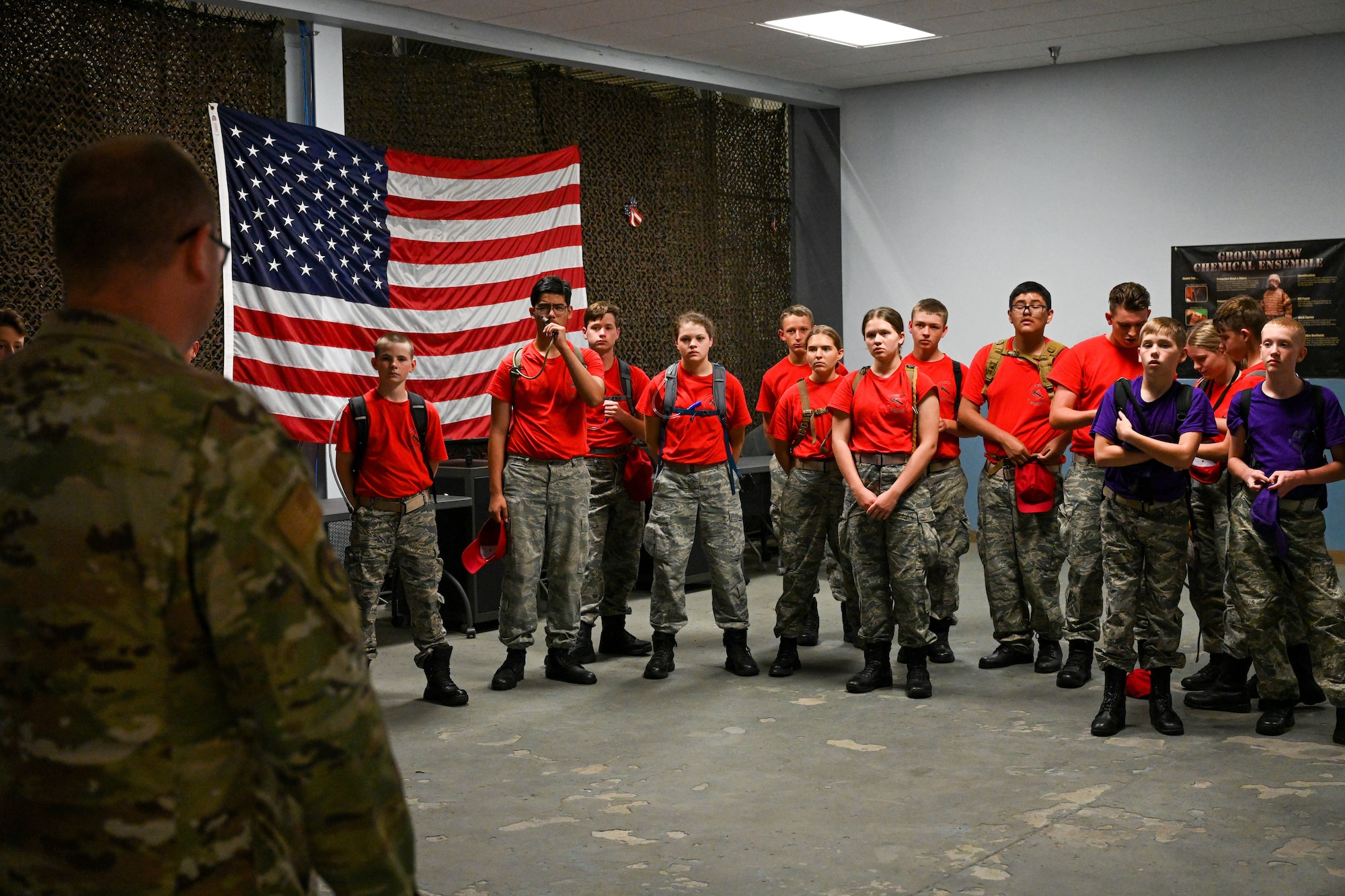 Civil Air Patrol Cadets wearing purple and red shirts listen to a briefing from a male Airman in OCPs.