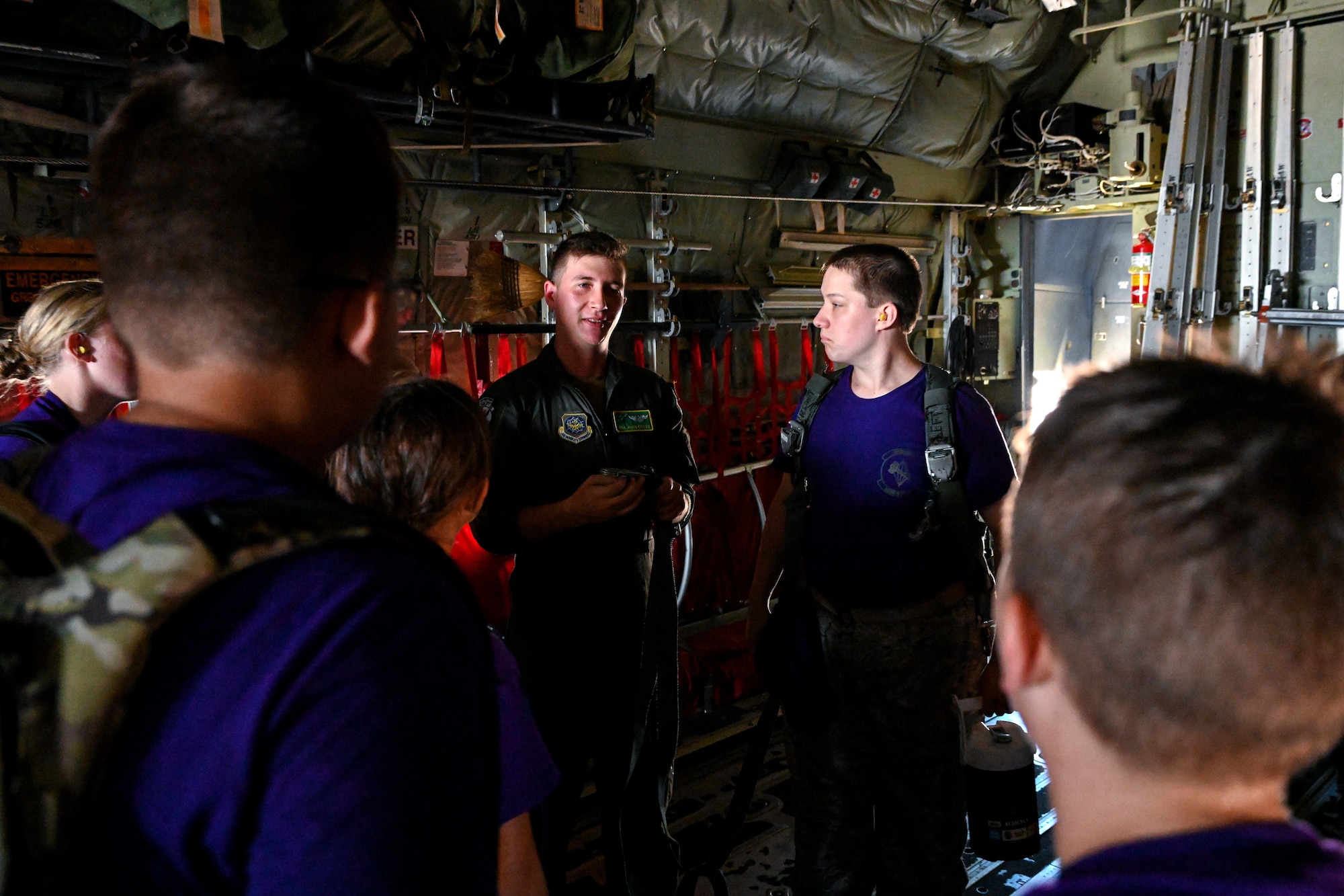 A male loadmaster wearing a flight suit speaks to Civil Air Patrol Cadets wearing purple shirts inside of a military aircraft.