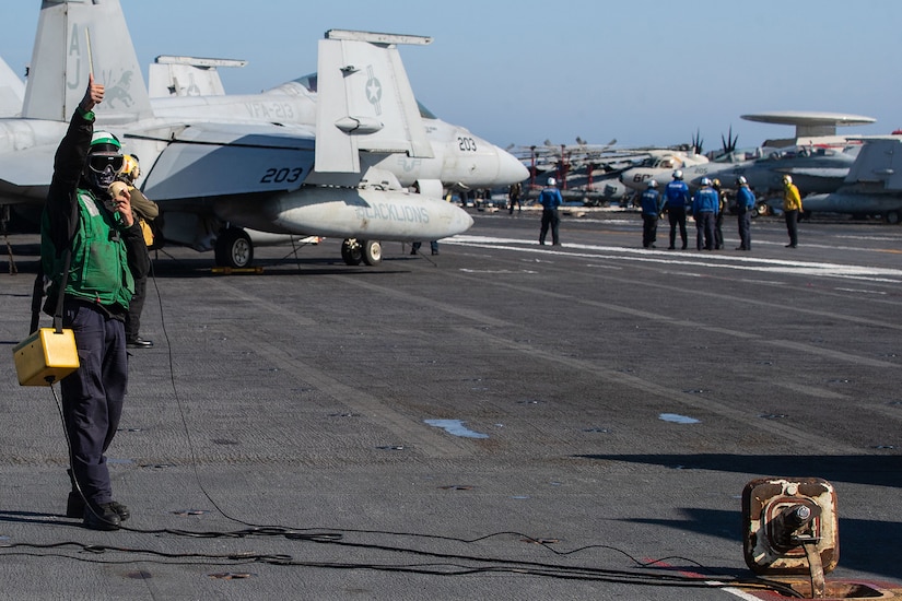 A service member wearing a green flight deck jersey gives a thumbs up hand signal.