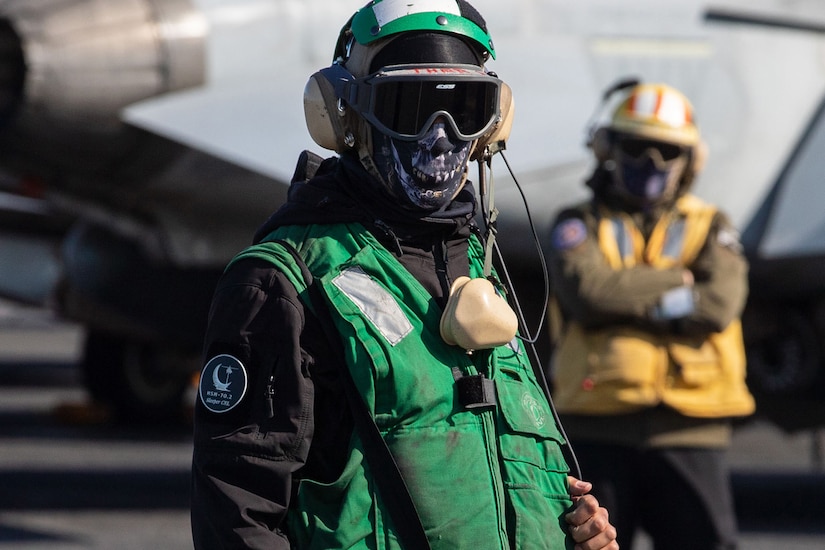 A service member wearing a green flight deck jersey stands on the flight deck in front of another service member in a yellow flight deck jersey.