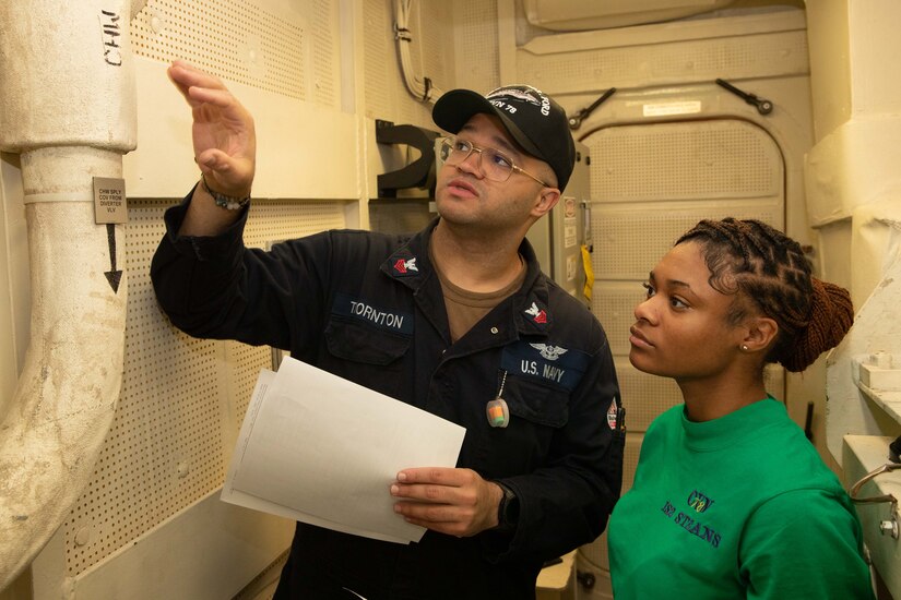 Two service members inspect a pipe with the lettering “CHW” on it.