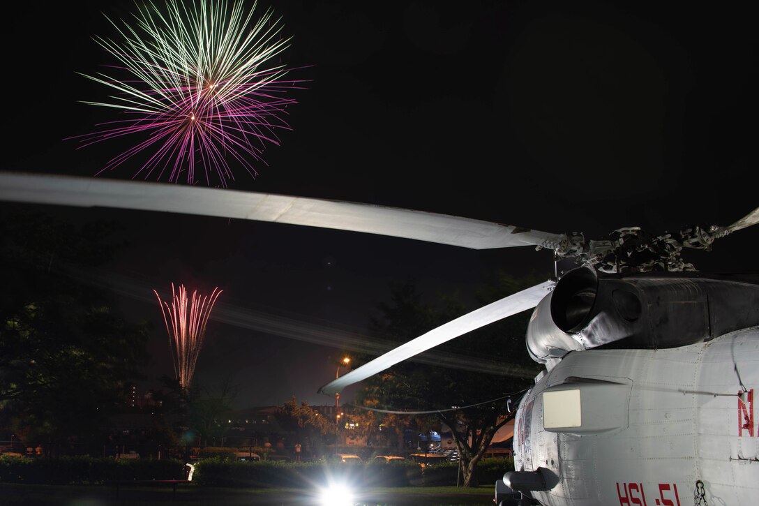 Fireworks adorn a dark sky above a field, with a helicopter parked in the foreground.