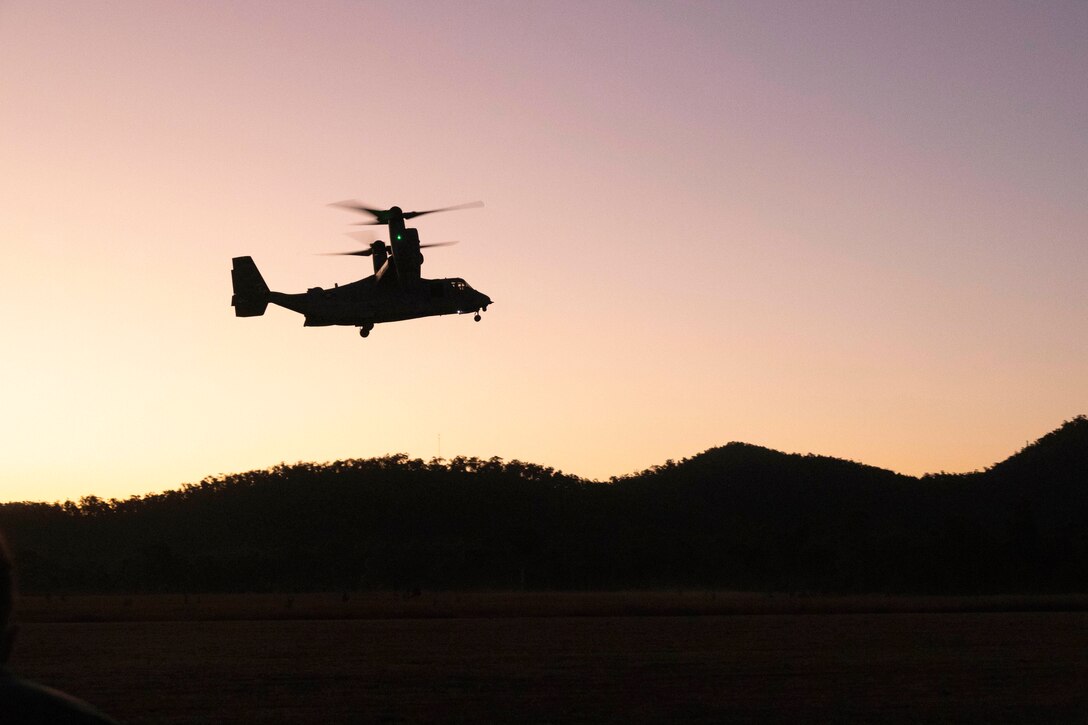 An aircraft flies over mountains under an orangish sky as shown in silhouette.