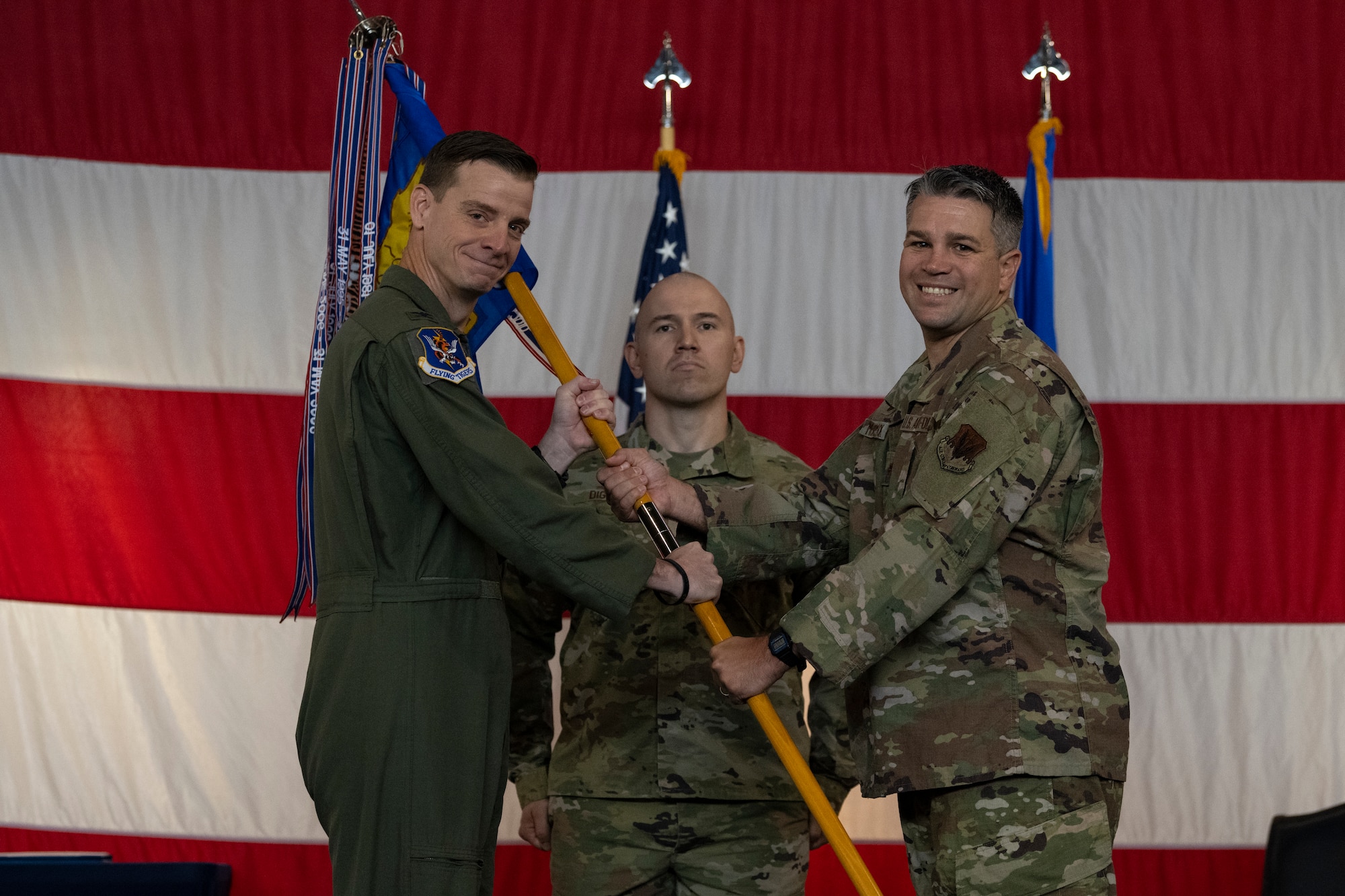 A photo of two U.S. Military uniformed Airmen holding a guidon between the both of them, smiling at the camera.