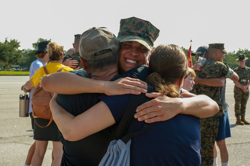 A Marine in uniform smiles as he hugs a man and woman.
