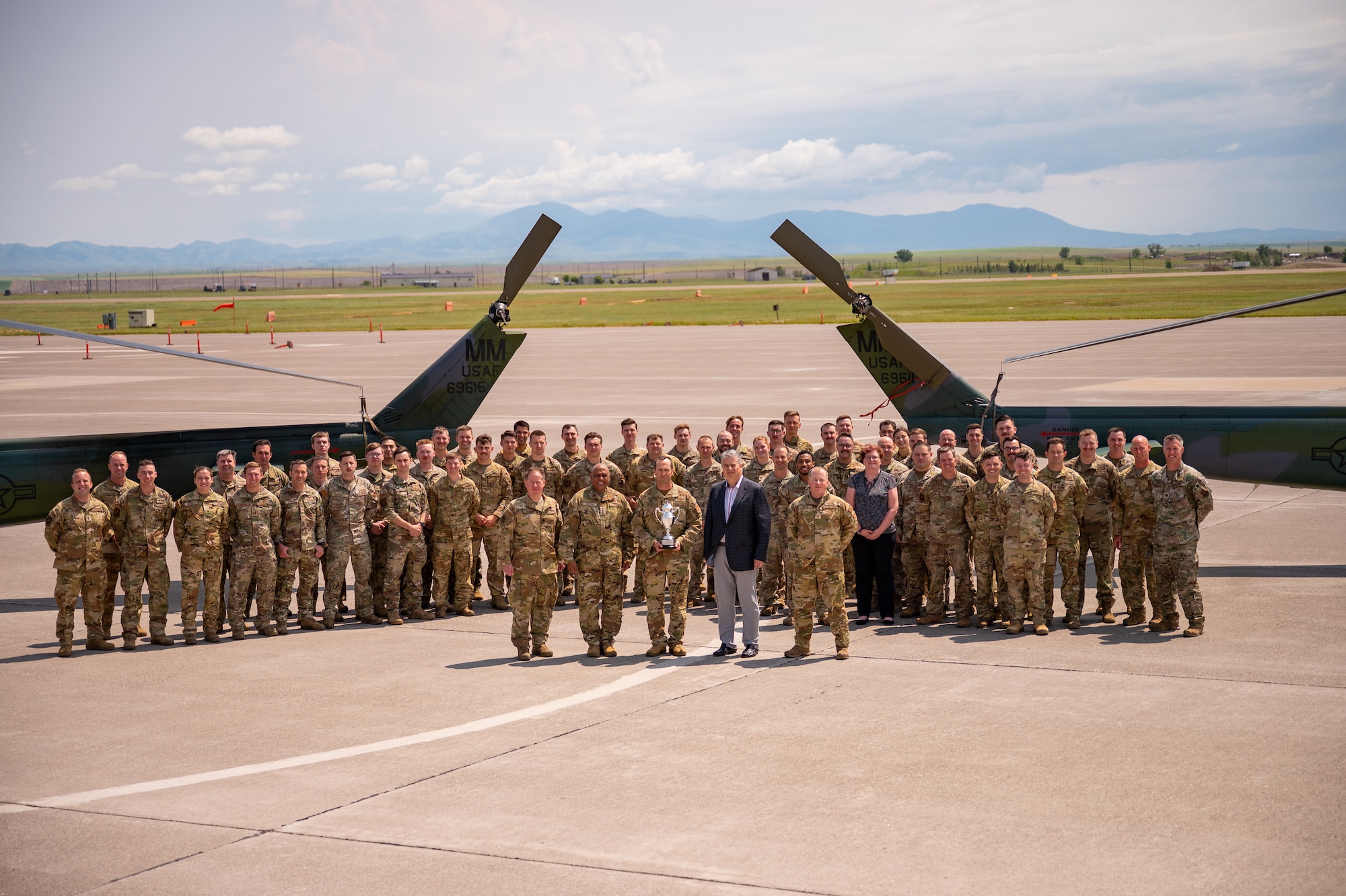 A large group of military members in uniform stand in formation in front of the tails of two helicopters.