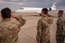 Three men in military uniform salute a plane as it taxis on a flightline in front of them.