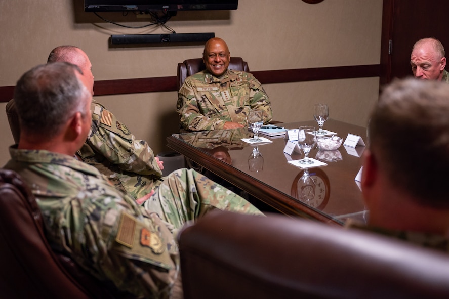 Men in military uniforms sit around a conference room table as they talk together.