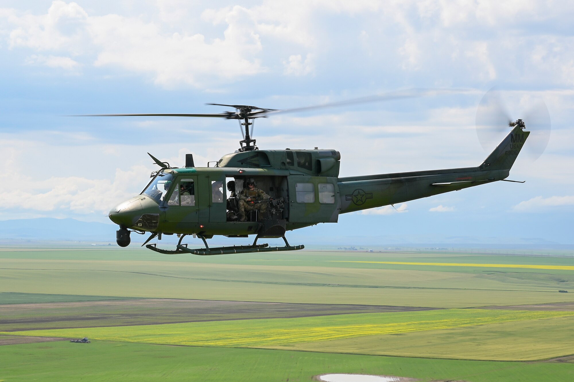 A UH-1N Huey helicopter flies against blue sky over green patches of farmland.