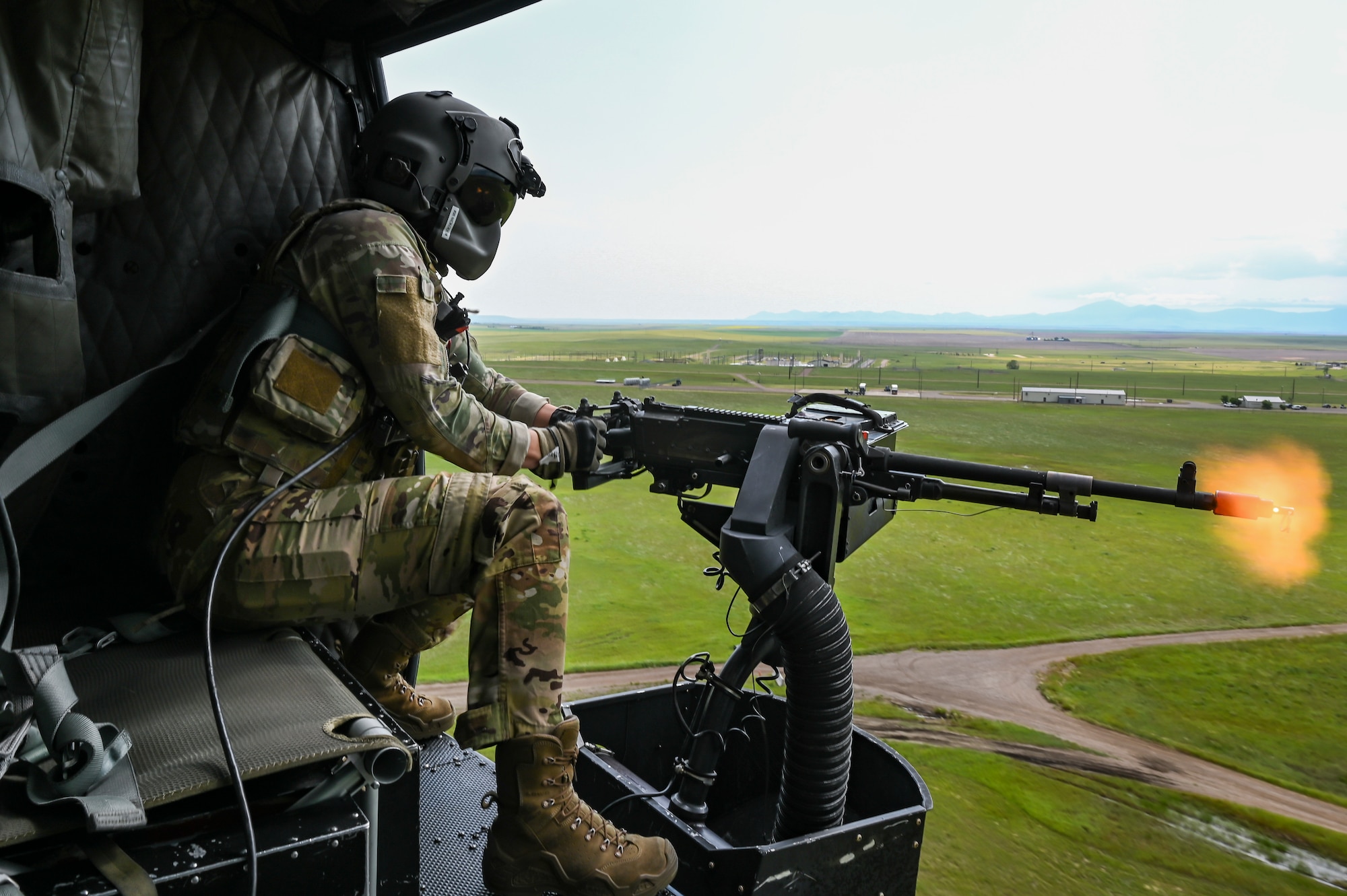 An Airman in uniform shoots blanks from an M240 machine gun out of a helicopter as it flies over green farmland.