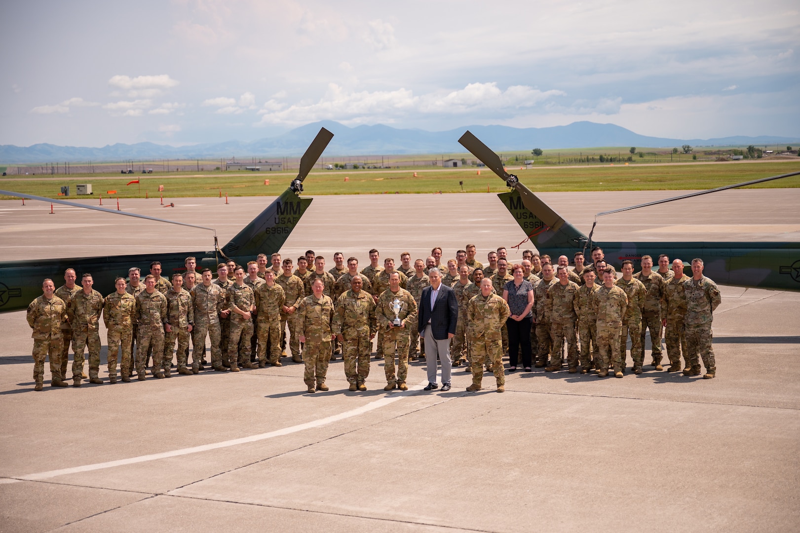 A large group of military members in uniform stand in formation in front of the tails of two helicopters.