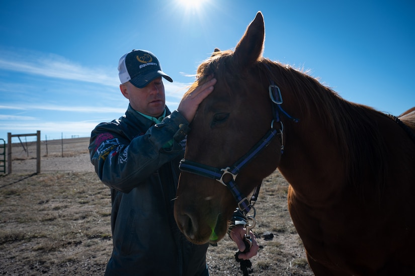 A man in a blue leather jacket filled with patches pets a horse in front of a farm.