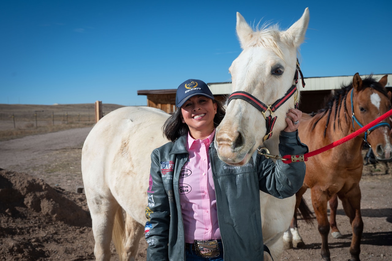 A woman in a blue leather jacket filled with patches poses in front of two horses.
