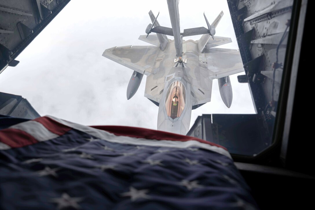 A military jet is refueled in midair as seen from above. A U.S. flag is in the foreground.