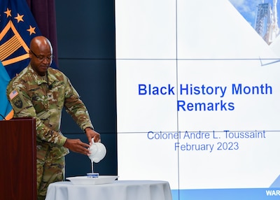 Uniformed soldier pours water from a tea pot into a tea cup on stage.