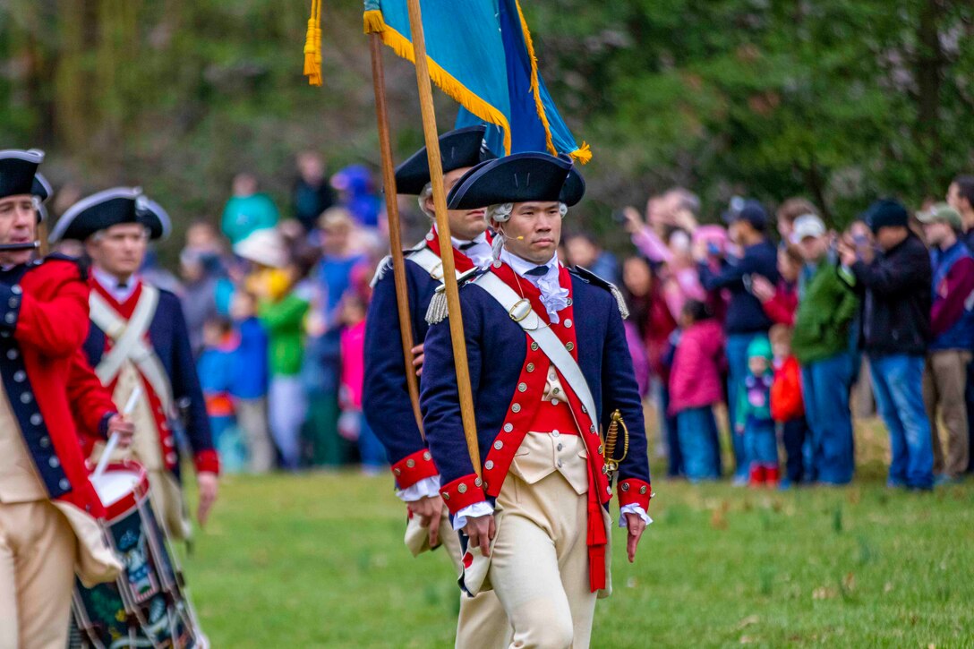 Soldiers  in period costume walk in formation; some hold flag poles and others play instruments.