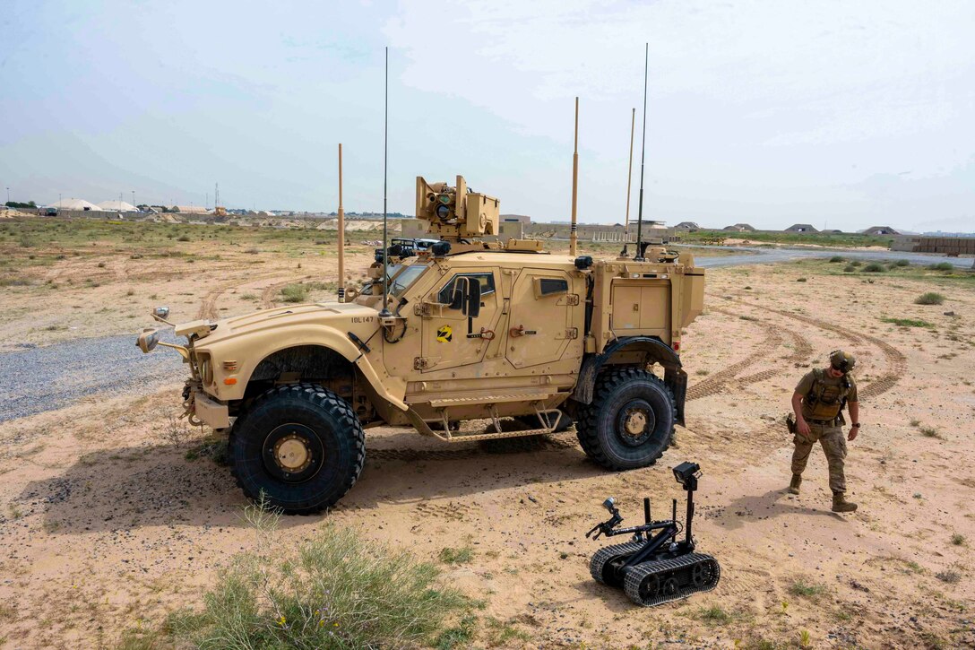 A soldier walks toward a small robot that sits on the ground next to a parked military vehicle.