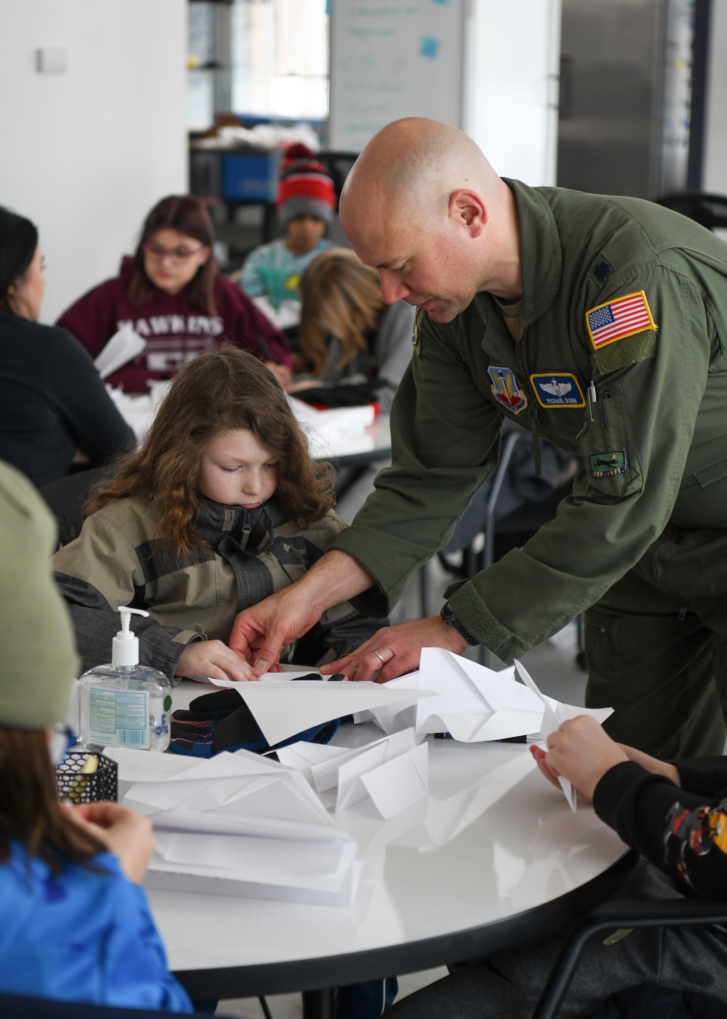 A man in a green uniform helps a child in a green coat with making a paper airplane