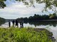 A group of six people stand near a river bank with trees in the background and vegetation in the foreground.