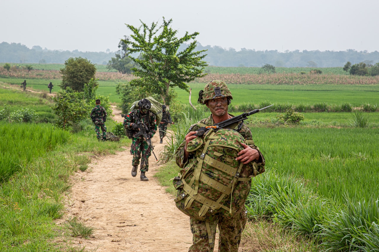 People in military uniform walk on a dirt path.