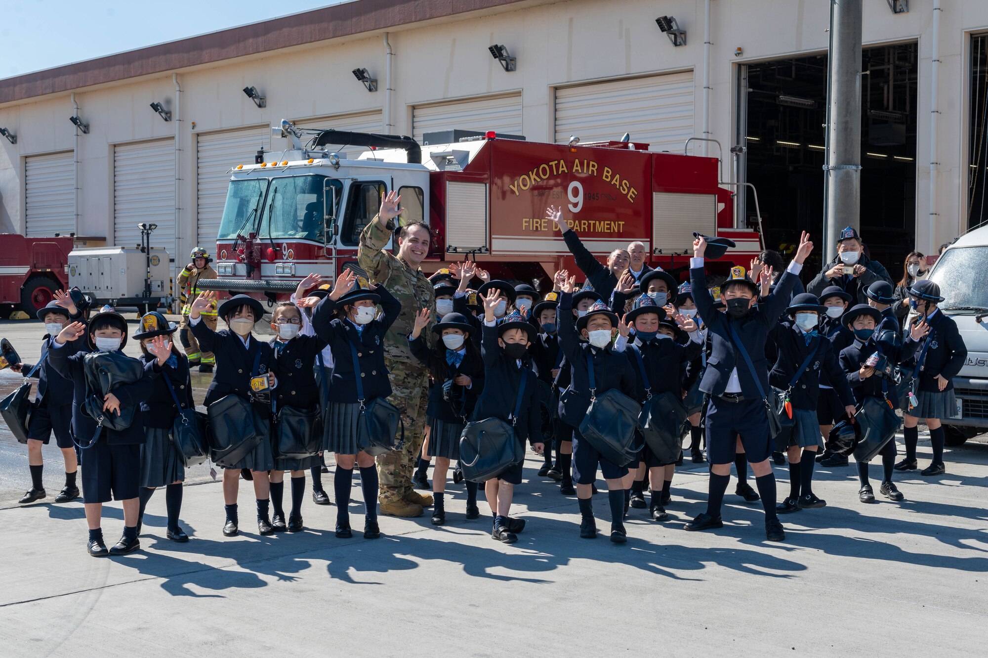 Standing outside in front of a fire truck, a white man and numerous Japanese children smile and wave to the camera.