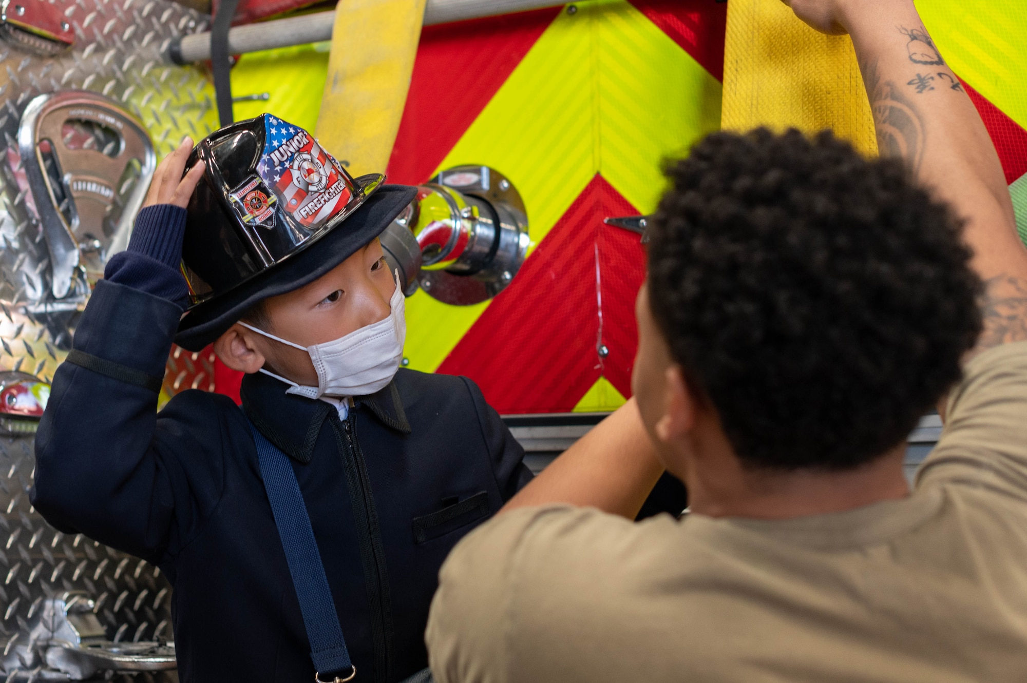 A Japanese child stands on a fire truck and looks at a black man talking to him.
