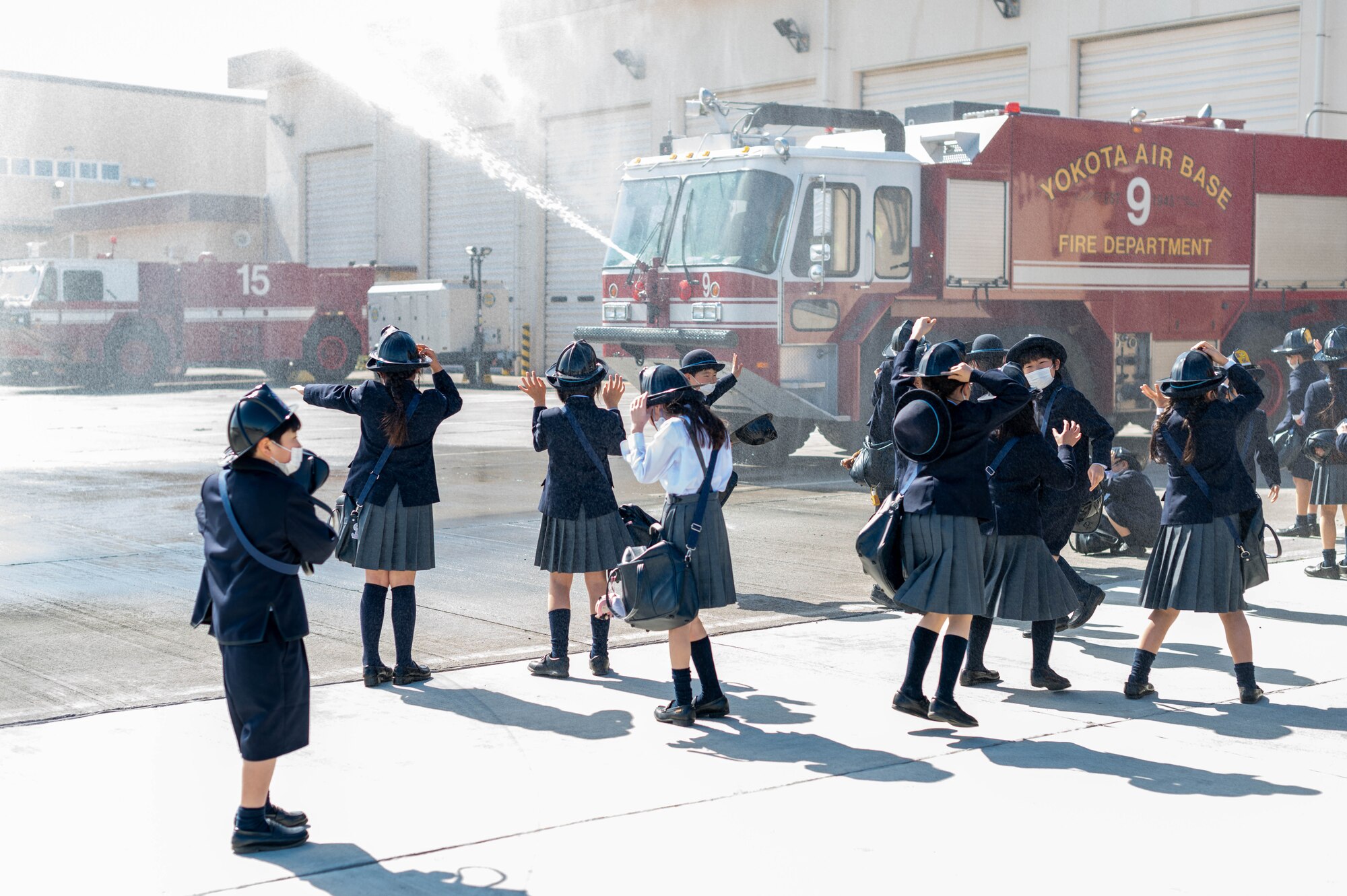 Numerous Japanese children smile, jump, and cover their heads as a fire truck sprays water in front of them.