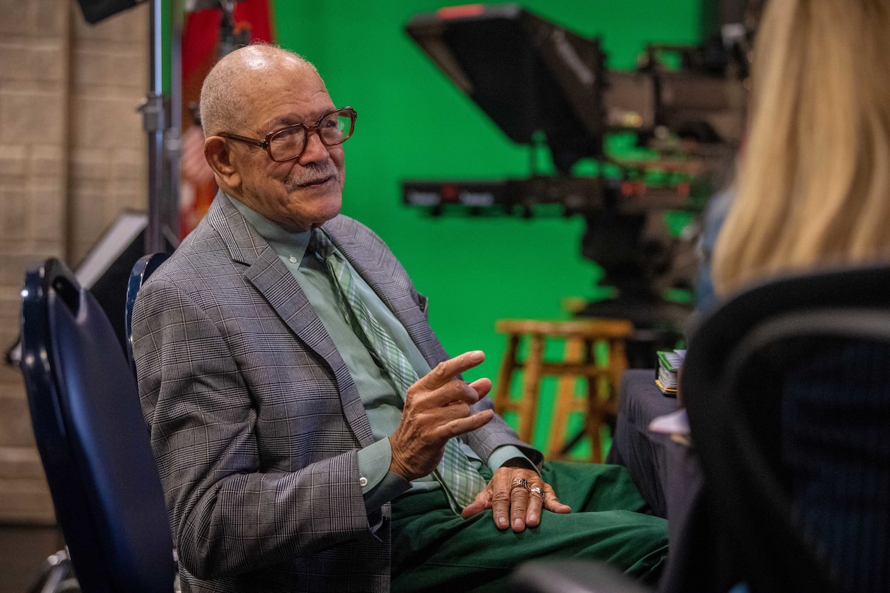 A veteran speaks to an interviewer on the sideline with a green screen and tv camera in the backdrop.