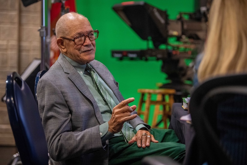 A veteran speaks to an interviewer on the sideline with a green screen and tv camera in the backdrop.