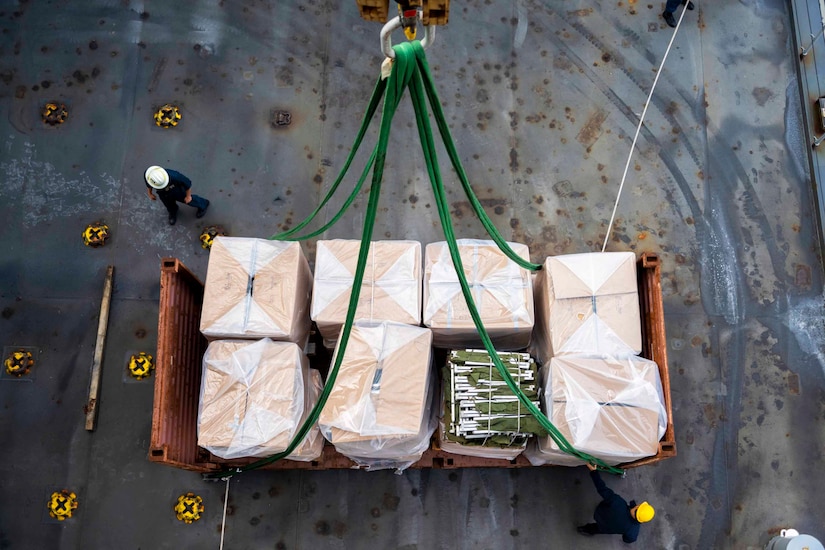 Navy and civilian personnel check the straps on a flat rack container.