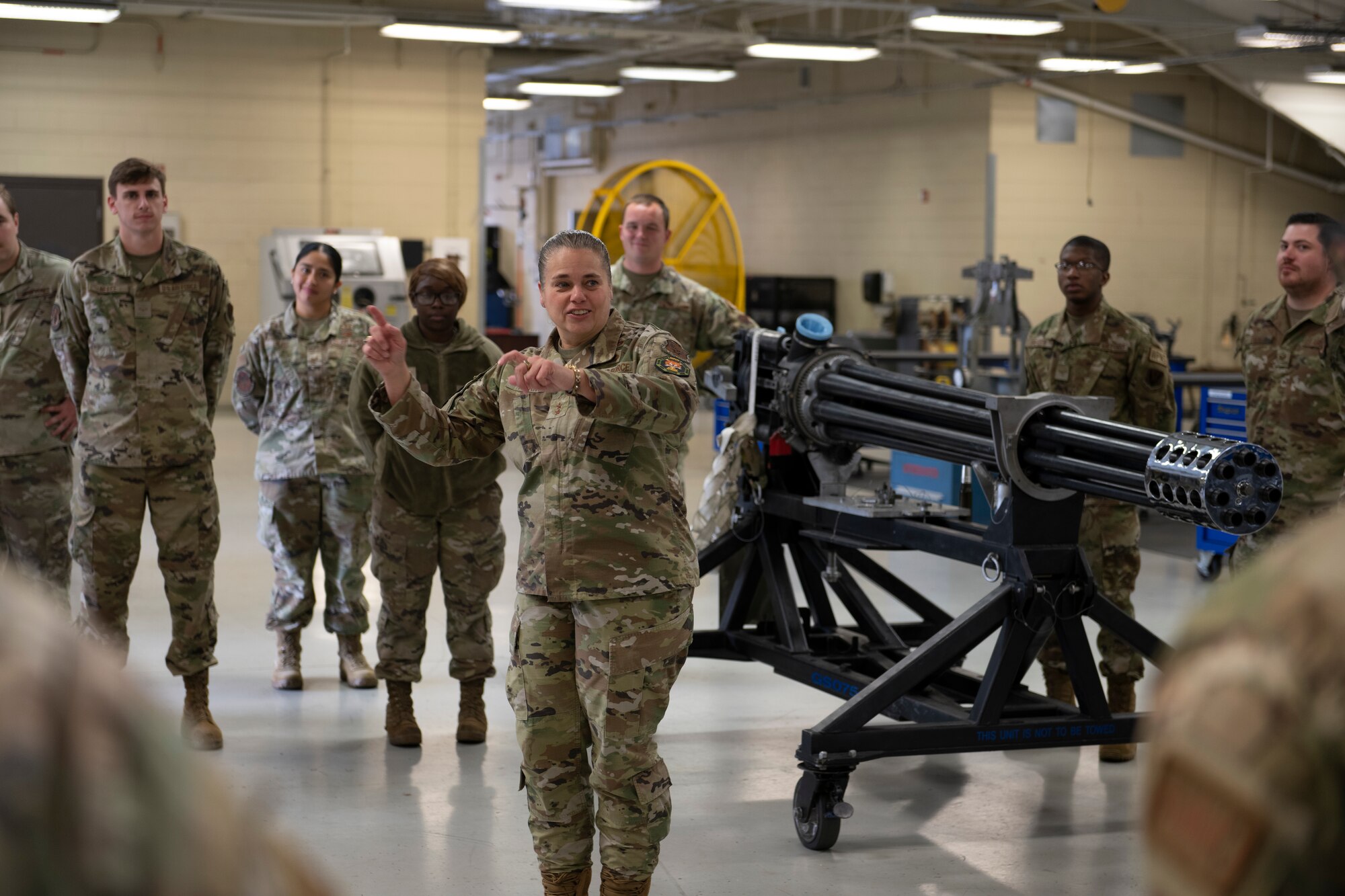 A photo of a woman standing in front of a weapons system with people standing in a circle around her, while she talks with her hands.