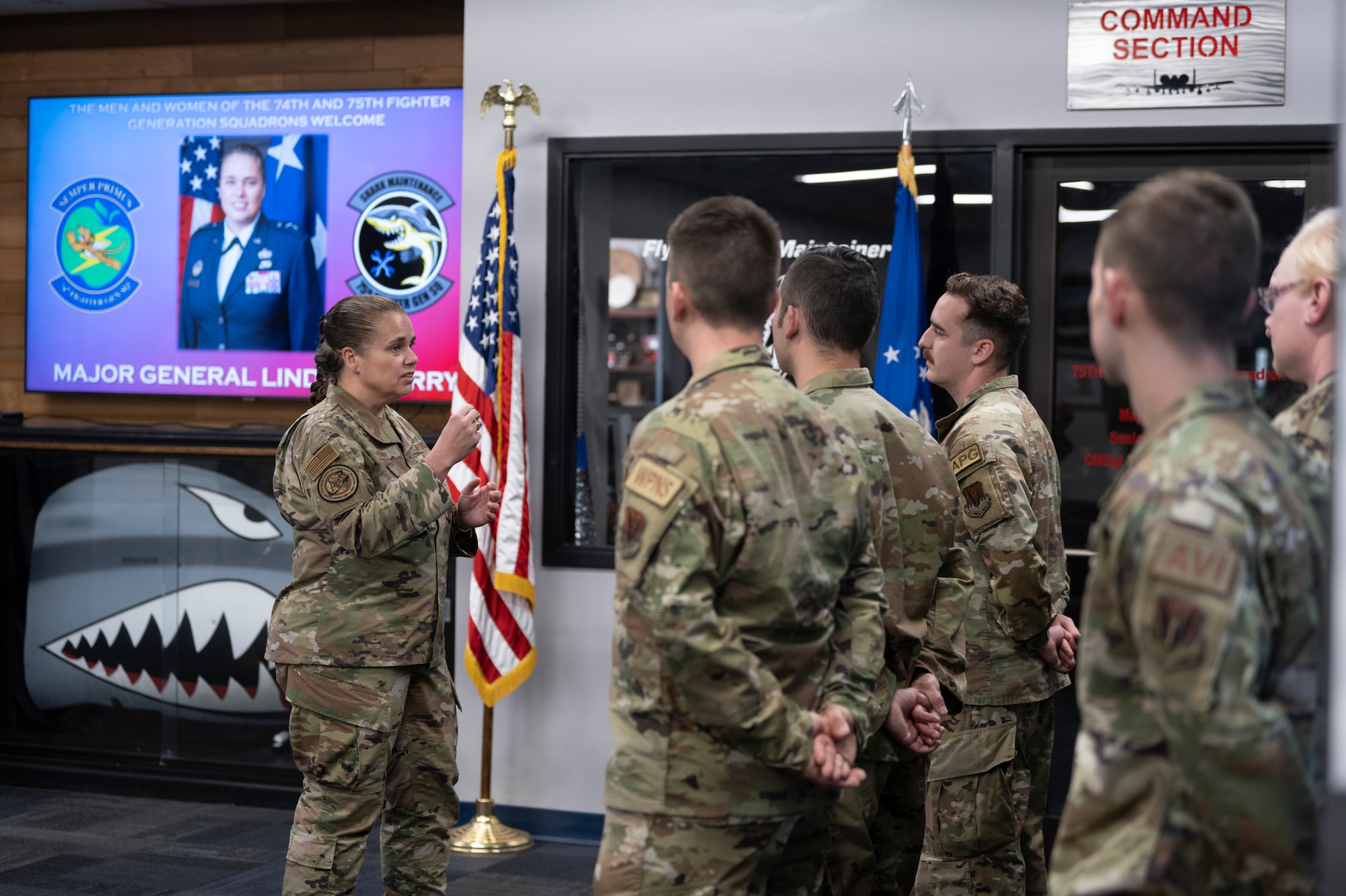 A photo of a woman on the left side talking to a group of people on the right side of the screen who are standing in a formation.