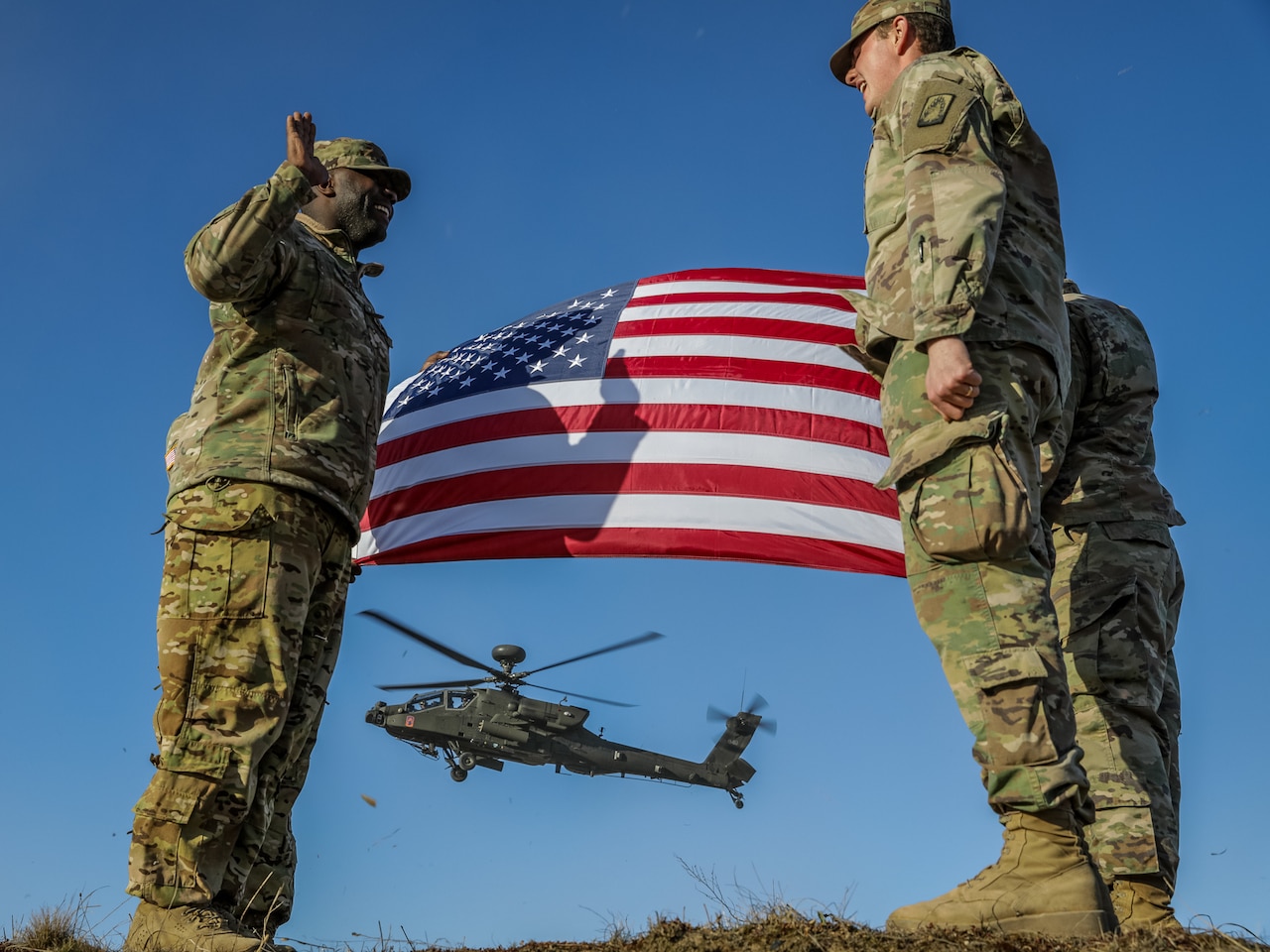 Three uniformed service members stand near one another. In the background are a U.S. flag and a helicopter.