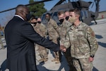 A man dressed in a business suit shakes hands with a soldier. There is a helicopter on the tarmac in the background.