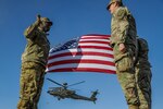 Three uniformed service members stand near one another. In the background are a U.S. flag and a helicopter.