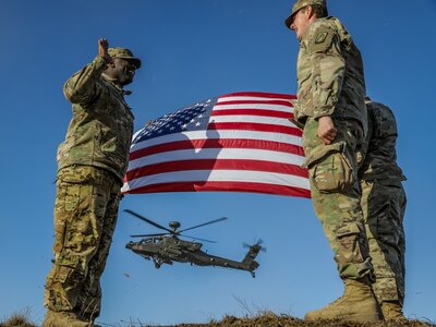 Three uniformed service members stand near one another. In the background are a U.S. flag and a helicopter.